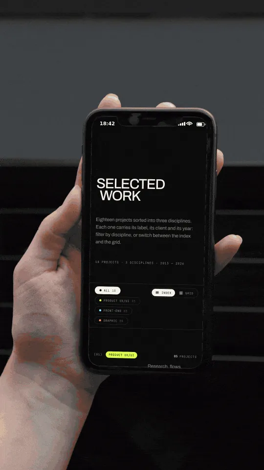 Photographic mockup: a hand holds a phone with the portfolio work archive scrolling, from the front-end development rows to the graphic design section